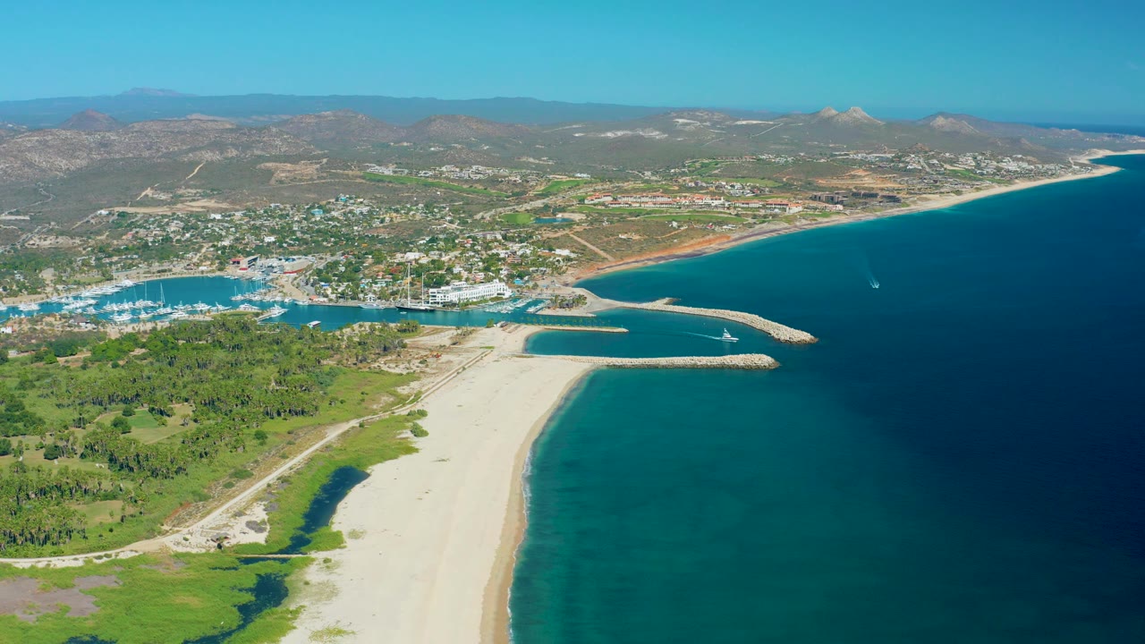 Wide San Jose del Cabo aerial showing the breakwater extending into blue water, marina boats clustered left of the inlet, a long beach, and no visible people.