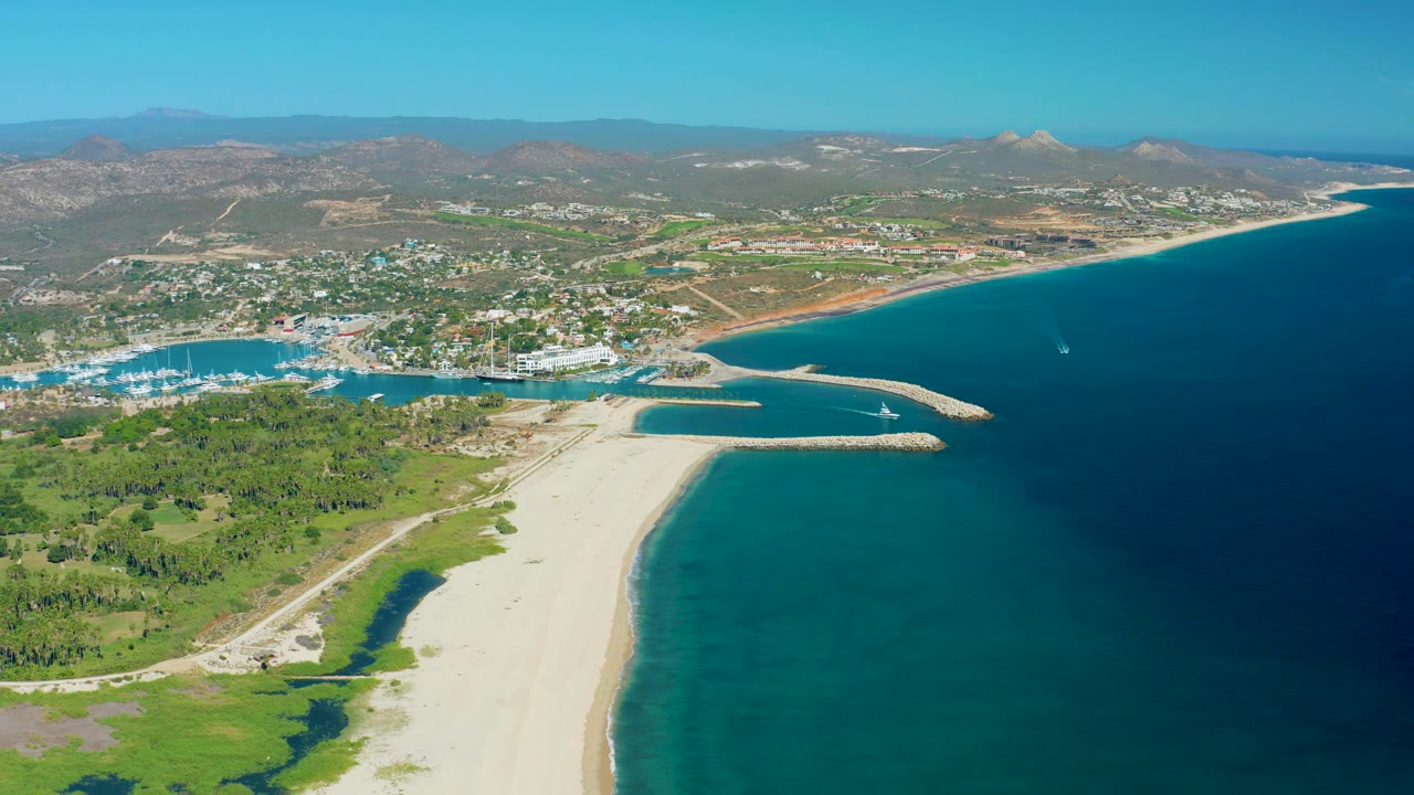 Aerial coastline view with marina, beach, ocean, green land, and hills.