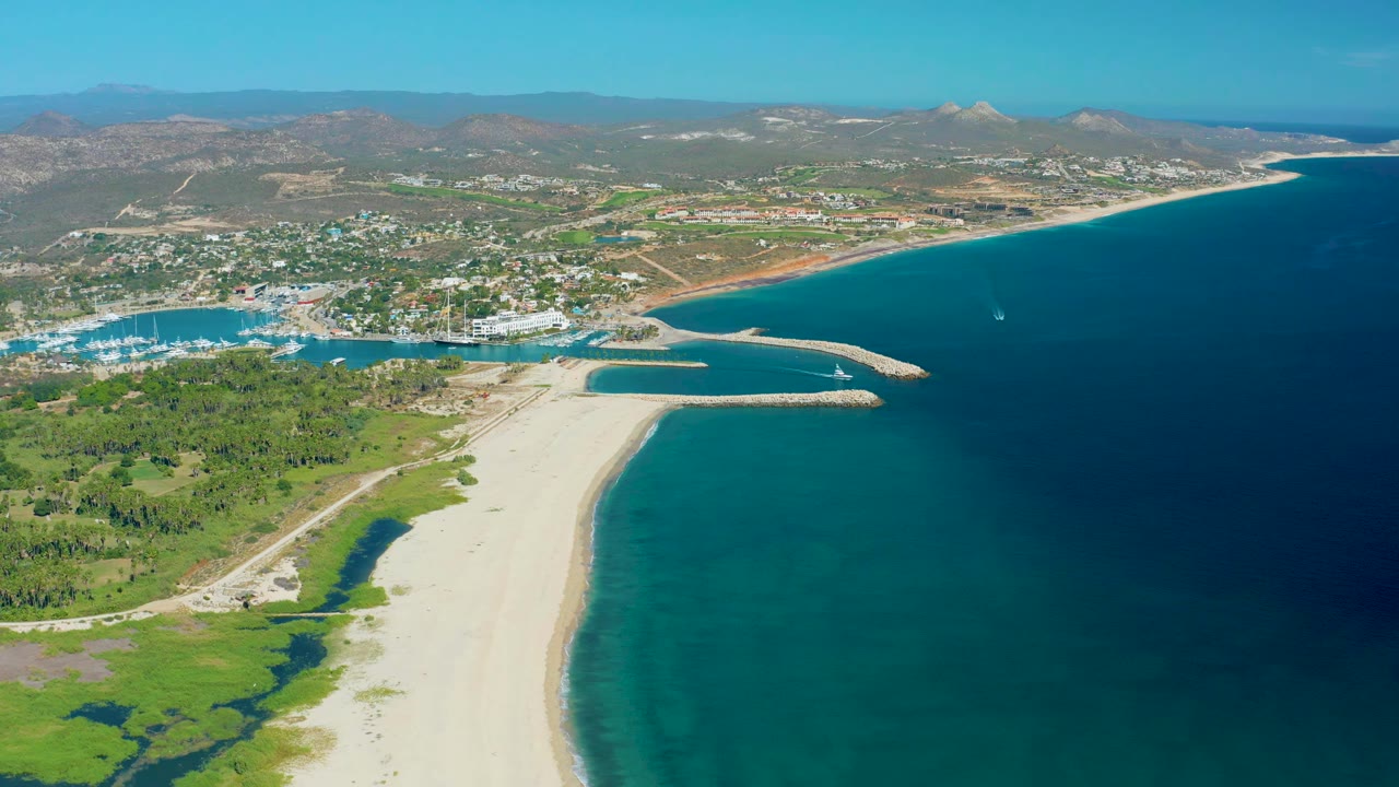 Aerial view in San Jose del Cabo with the sandbar running toward the harbor entrance, boats clustered in the marina, green lagoon vegetation at left, blue ocean at right, and no visible people.