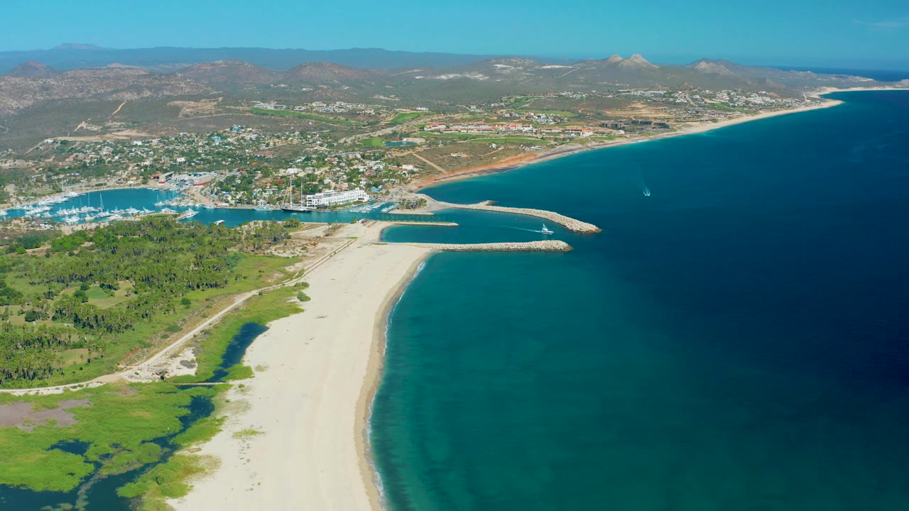 Wide aerial frame of San Jose del Cabo with a bright beach in the lower frame, marina and town across the middle distance, arid hills beyond, open sea to the right, and no visible people.