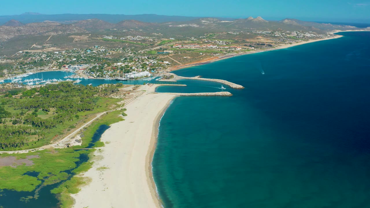San Jose del Cabo aerial still with lagoon greenery and pale sand in the foreground, a harbor with boats at left, a curved coastline and deep blue sea at right, and no visible people.