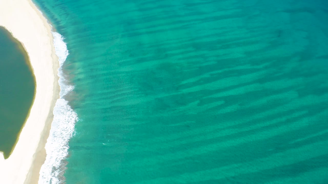 Overhead San Jose del Cabo beach view with bright sand on the left, a thin band of breaking foam, and green-blue water with visible tonal bands, with no visible people.