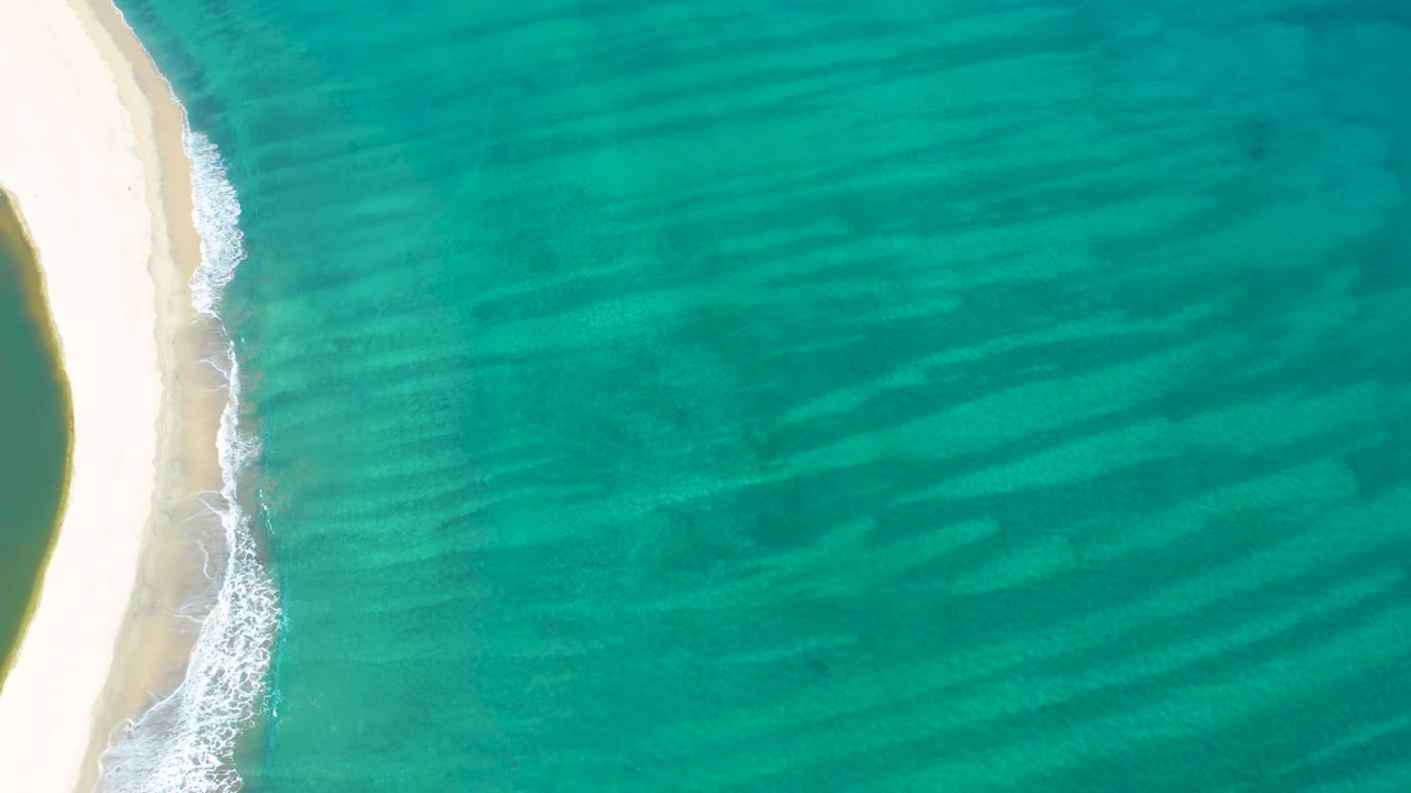 Aerial frame from San Jose del Cabo with the sandy beach pressed against the left side and clear blue-green water spreading across the frame, with no visible people.
