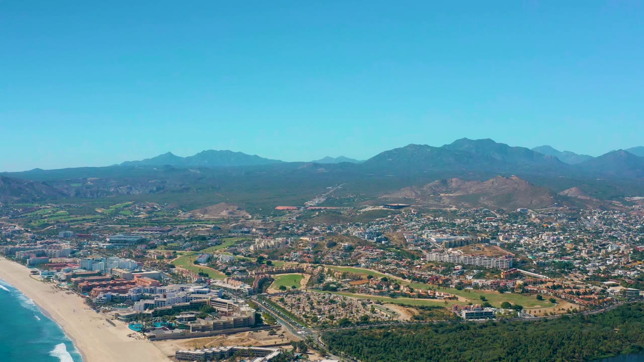 Early aerial view of San Jose Del Cabo with the beach curving along the left side, urban buildings and roads inland, green patches in the midground, mountain ridges behind, and no visible people.