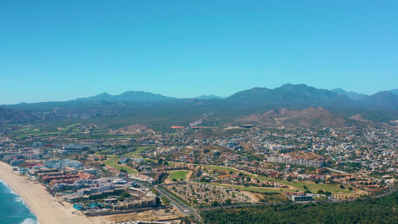 Aerial view over San Jose del Cabo showing blue ocean and sandy beach at left, coastal buildings and green open space in the center, dry hills and mountains beyond, with no visible people.