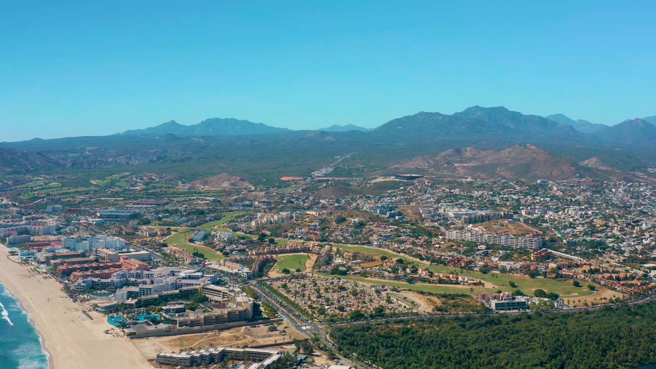 Aerial coastal city view with beach, roads, green areas, and mountain backdrop.