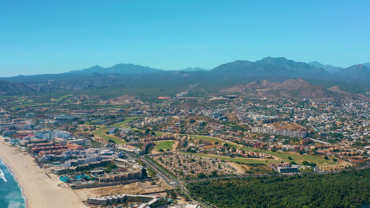 Mid-clip aerial frame of San Jose del Cabo with a long sandy beach on the left, resort-scale and residential buildings in the midground, patches of greenery, distant mountain ridges, and no visible people.