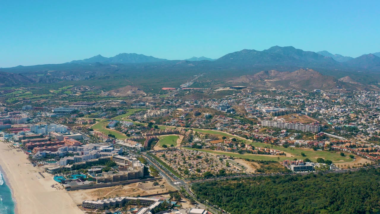 Wide aerial view over San Jose del Cabo with sandy beach and ocean at the lower left, city buildings and roads through the center, green fields beyond, distant mountains, and no visible people.