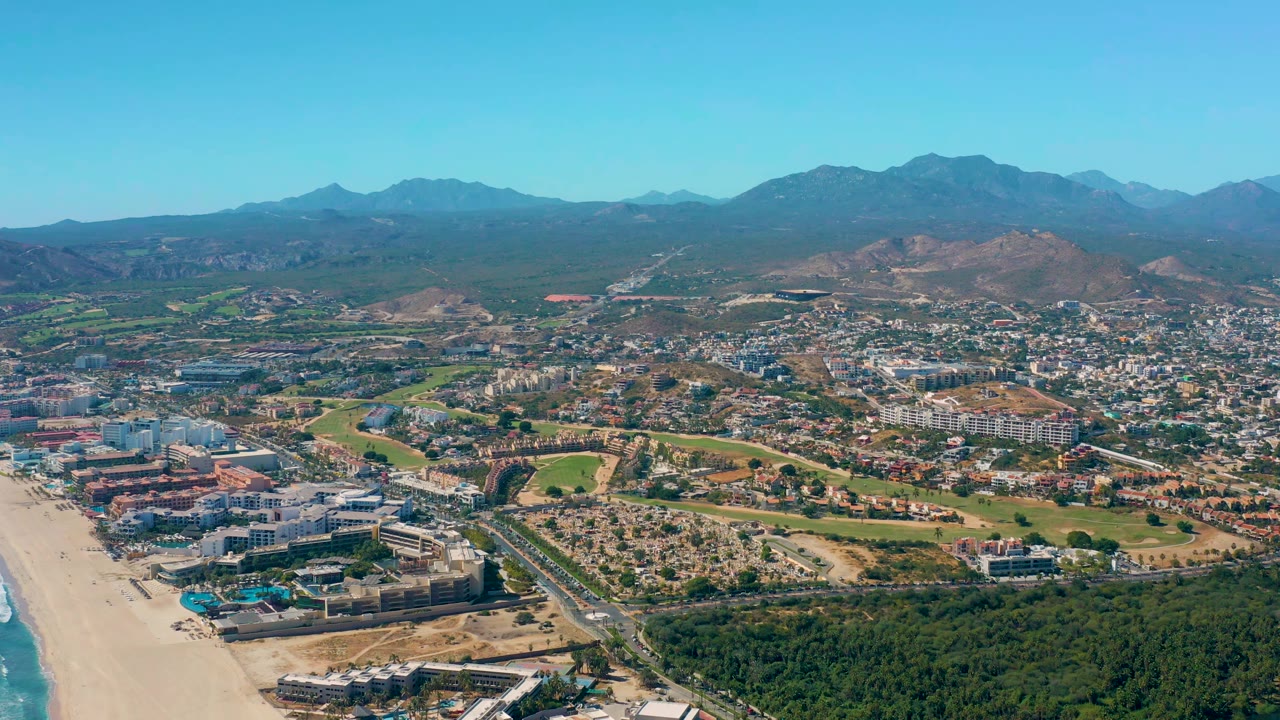 San Jose del Cabo aerial frame with the beach running along the left side, blue water at the edge, clustered beachfront buildings, a road corridor, inland neighborhoods, and no visible people.