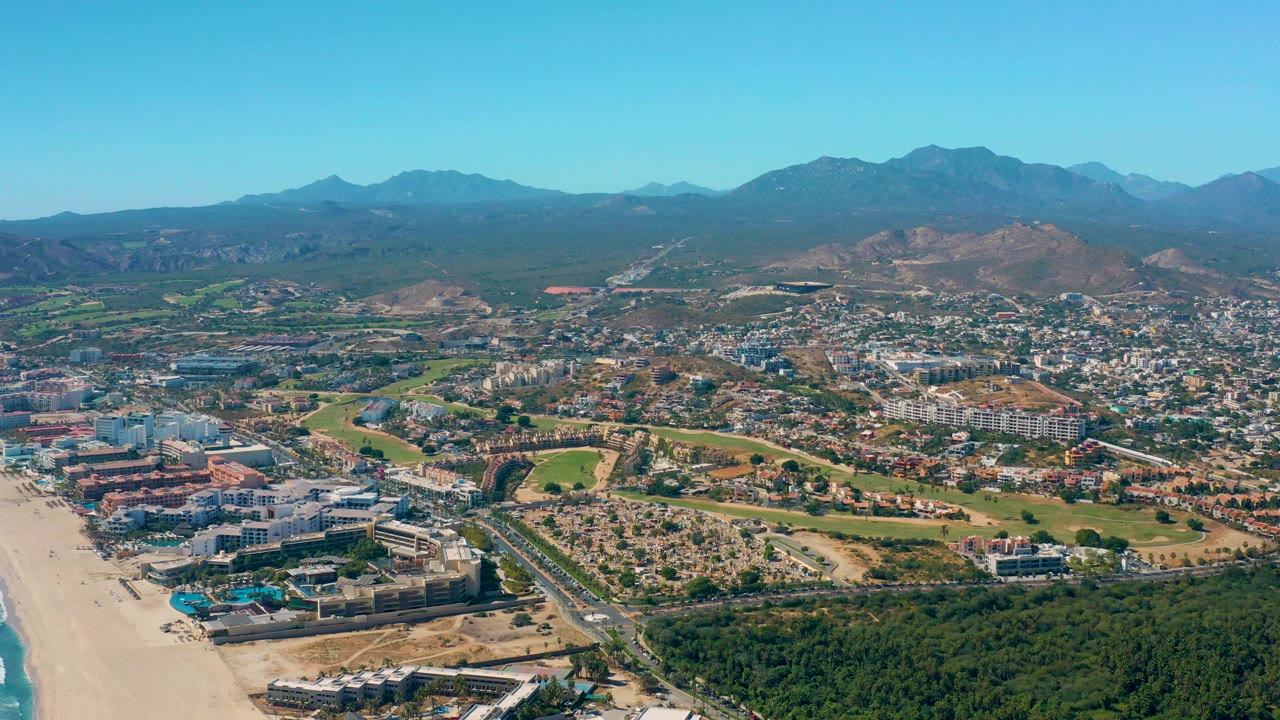Aerial panorama of beach, city streets, green areas, and distant mountains.