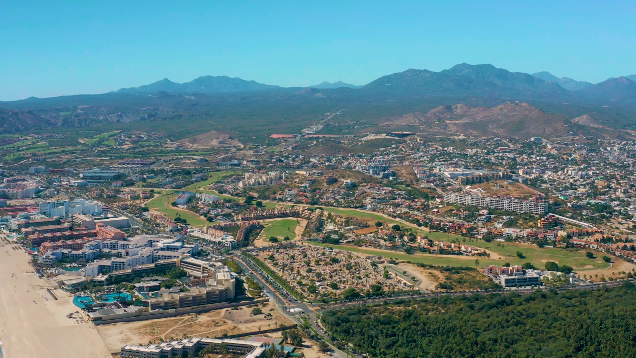 San Jose Del Cabo aerial frame with the beach still visible along the left edge, low buildings in the foreground, a road grid and town center in the middle, and no visible people.