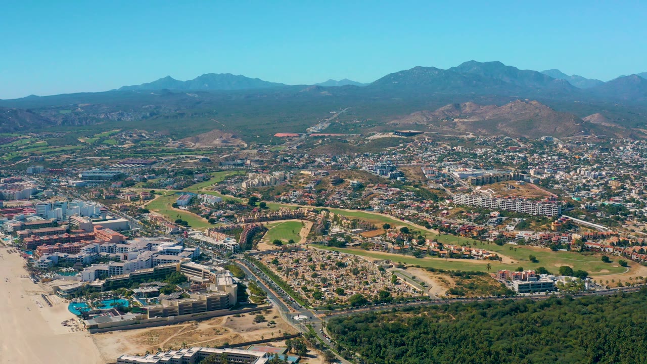 Mid-clip aerial frame of San Jose Del Cabo showing a broad sweep from beach and foreground buildings to dense town blocks, dry hills, blue mountain ridges, and no visible people.