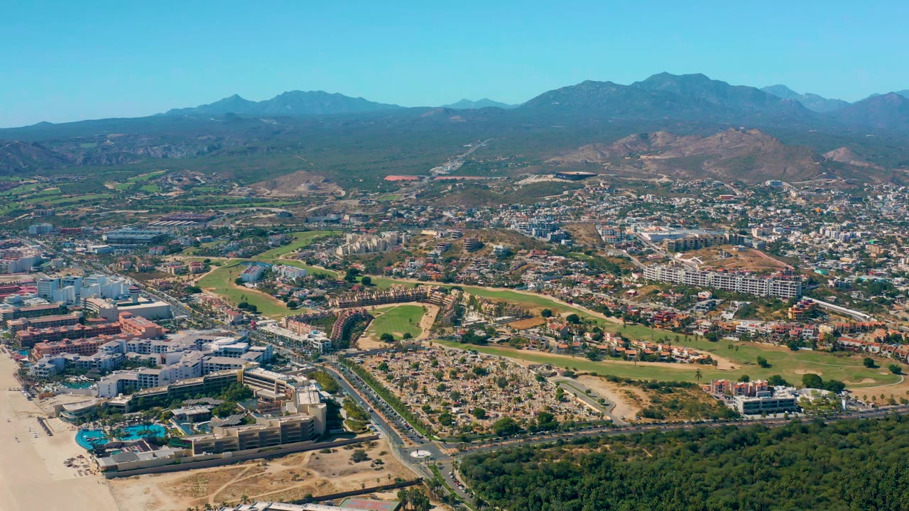 High aerial cityscape with beach edge, foreground road, buildings, green areas, and mountains.