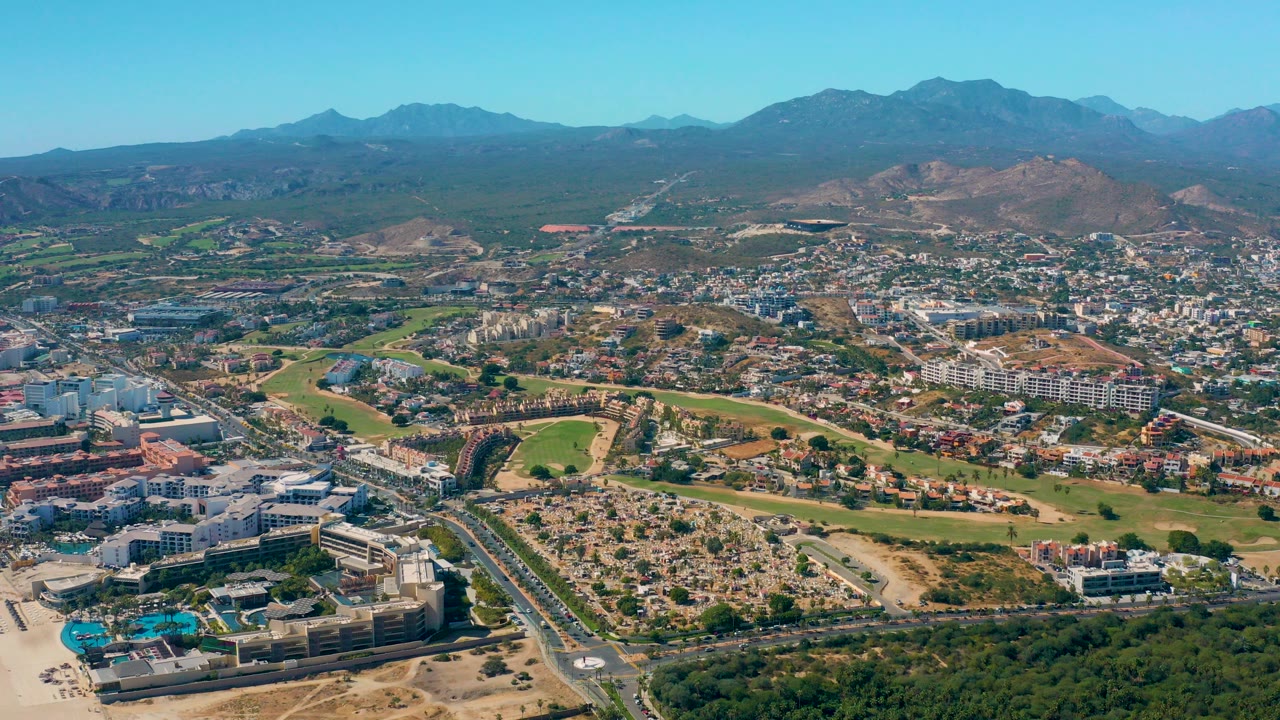 Opening aerial frame over San Jose del Cabo, Mexico, with a road and hotel-like buildings in the foreground, green fields and residential blocks beyond, mountains on the horizon, and no visible people.