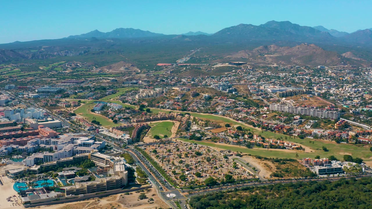 Aerial city edge with roads, houses, green fields, dry lots, and mountains
