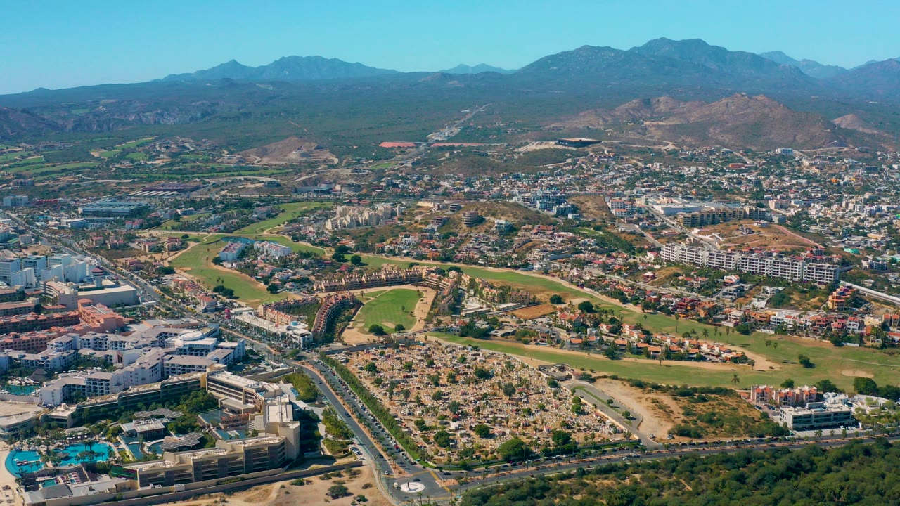 Wide aerial view over San Jose del Cabo with a divided road in the lower foreground, green fields and buildings through the center, distant hills and mountains, and no visible people.