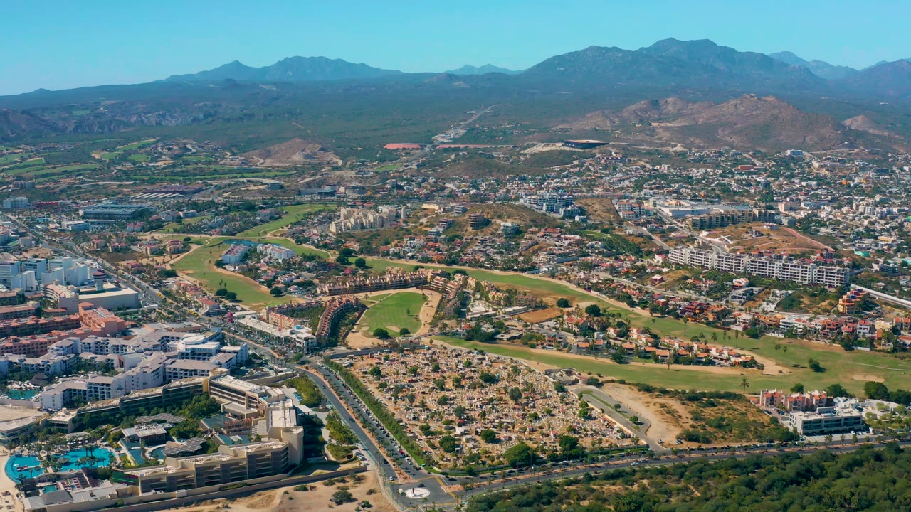 High drone-style view of San Jose del Cabo showing neighborhoods and commercial blocks around green fields, with dry hills and blue mountains across the horizon and no visible people.