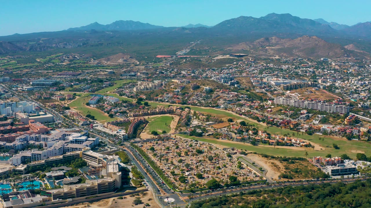 Aerial view of buildings, green fields, roads, dry hills, and mountain backdrop.