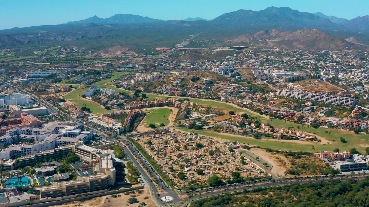 Opening aerial frame over San Jose Del Cabo with foreground roads and buildings, golf course greens through the center, dry hills, distant mountains, clear sky, and no visible people.