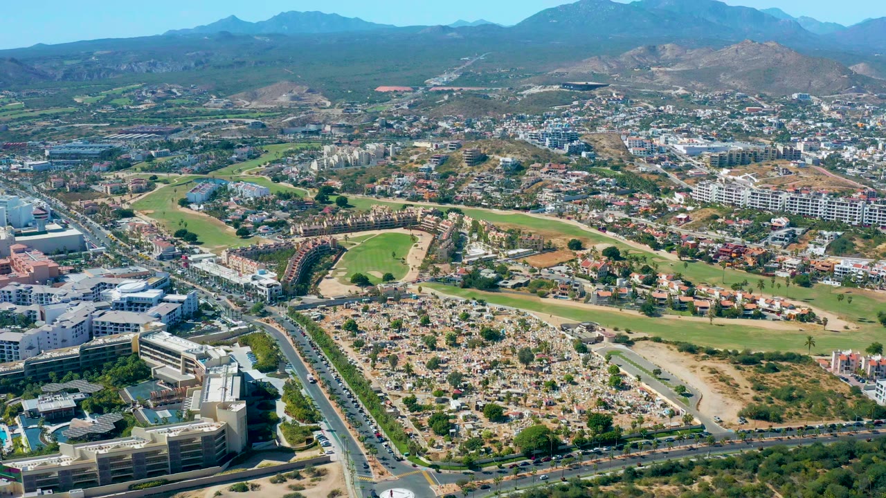 Opening aerial frame over San Jose Del Cabo with multilane roads in the foreground, a golf course and residential blocks in the middle distance, mountains on the horizon, and no visible people.