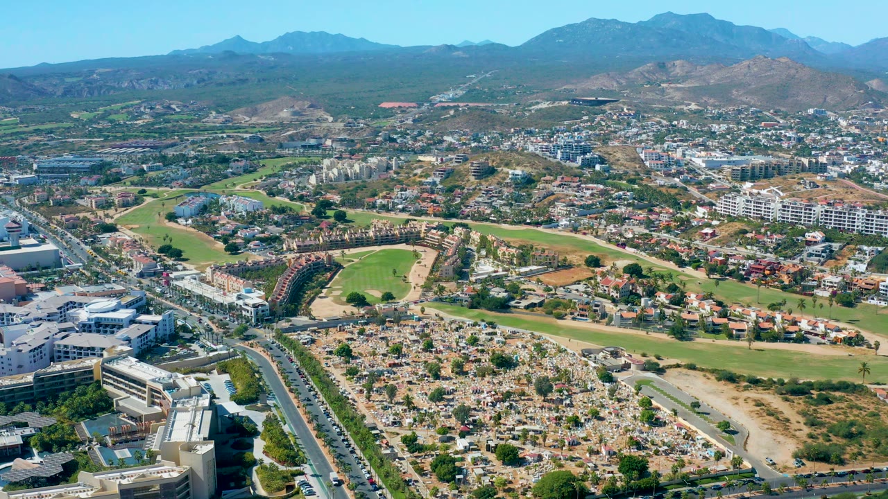 Wide aerial city view with traffic, homes, green areas, and hills.
