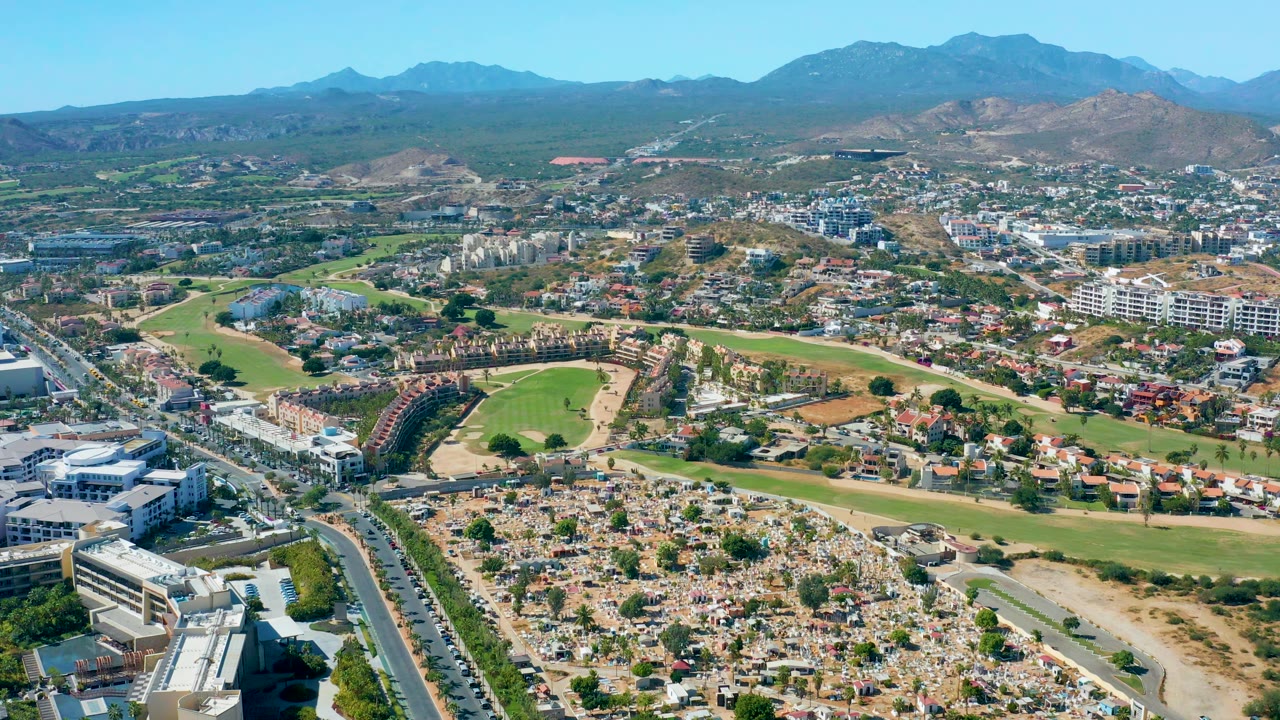 Aerial view of San Jose del Cabo one second into the clip, with a broad road corridor at left, dense low-rise buildings, green fairways, distant mountains, and no visible people.