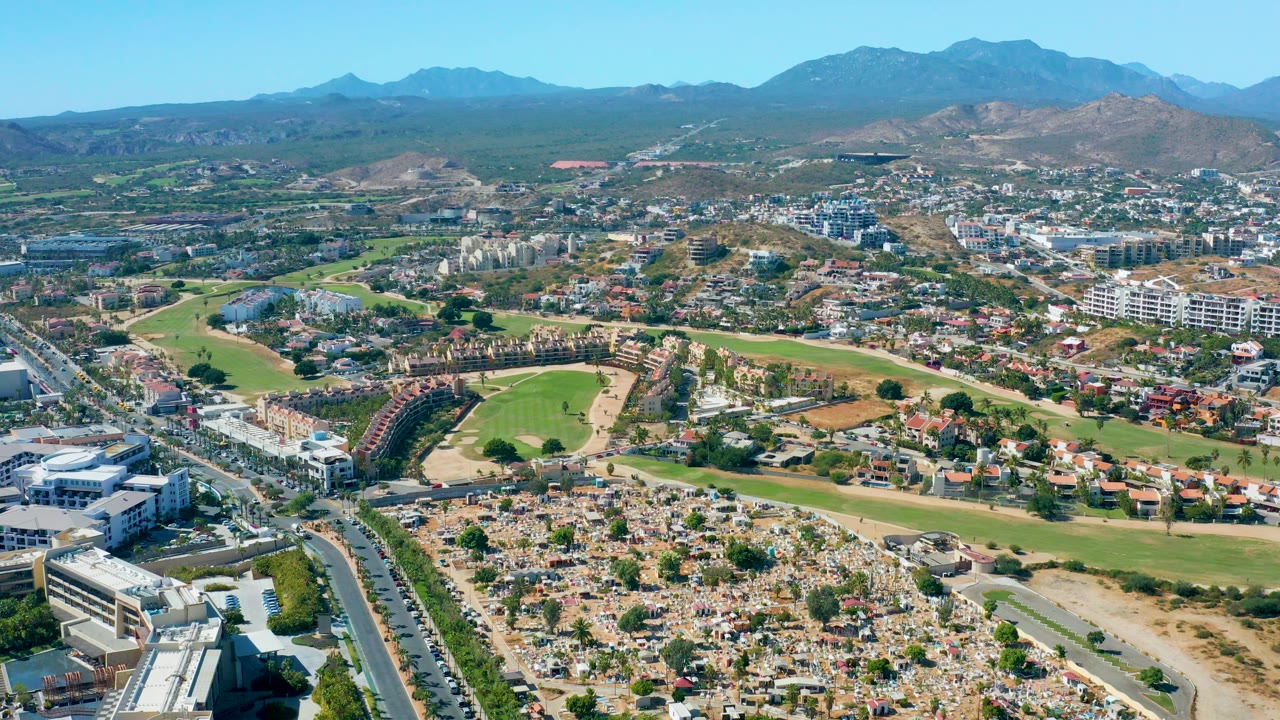 Aerial pan over a golf course edge, roads, buildings, and distant mountains.