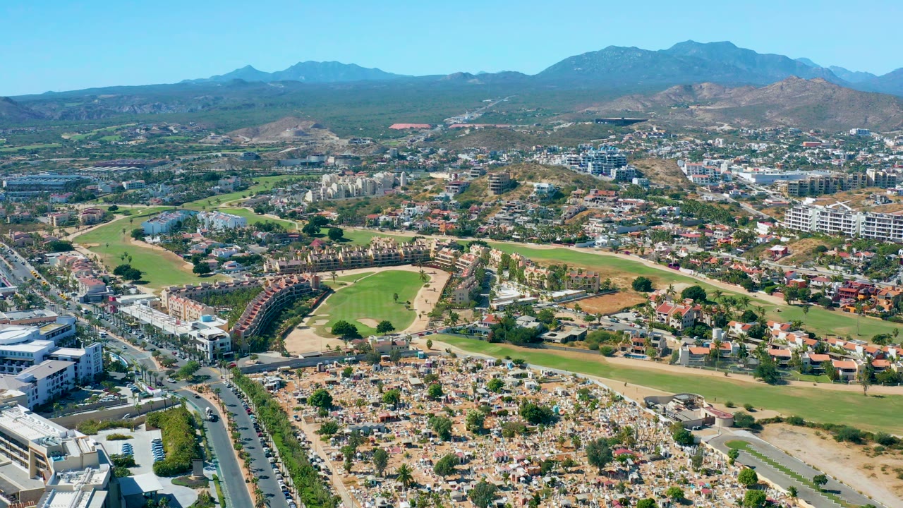 Aerial frame in San Jose del Cabo showing more of the golf course fairways across the midground, with houses, a busy road, cemetery-like foreground rows, mountains, and no visible people.