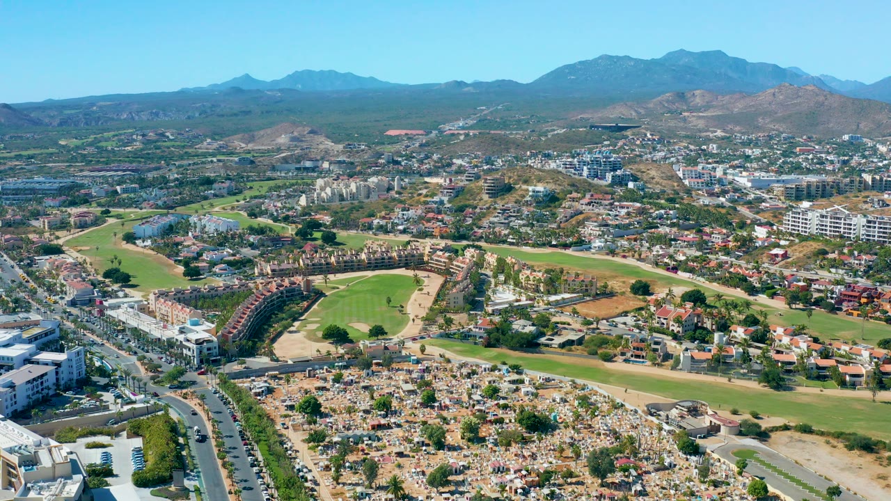 Aerial neighborhood scene with fairways, traffic, buildings, cemetery-like foreground, and mountains
