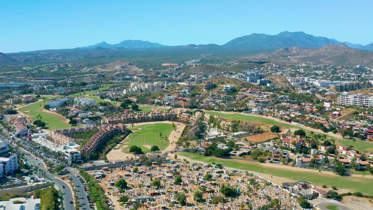 Wide aerial frame of San Jose del Cabo showing suburban homes, curving roads, green fields, distant mountains, clear blue sky, and no visible people.