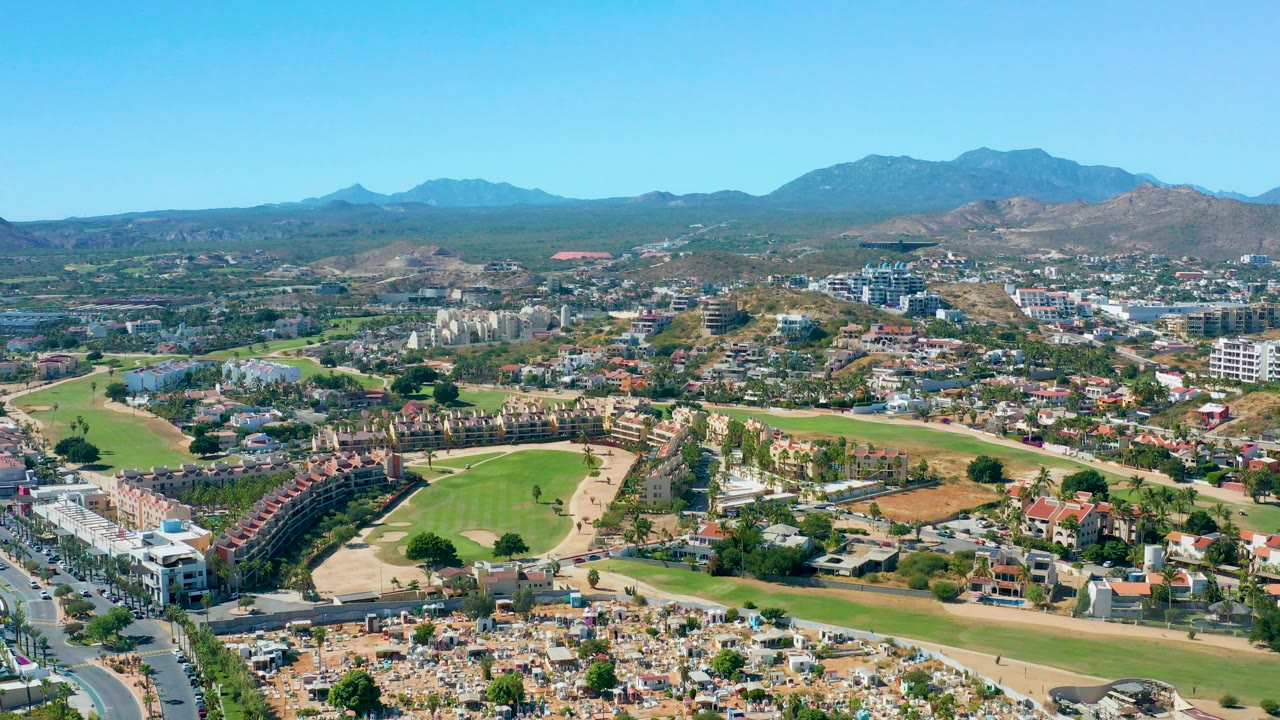 Wide aerial frame over San Jose del Cabo with red-roofed homes, green fairways, dry lots, roads, distant mountains, clear blue sky, and no visible people.