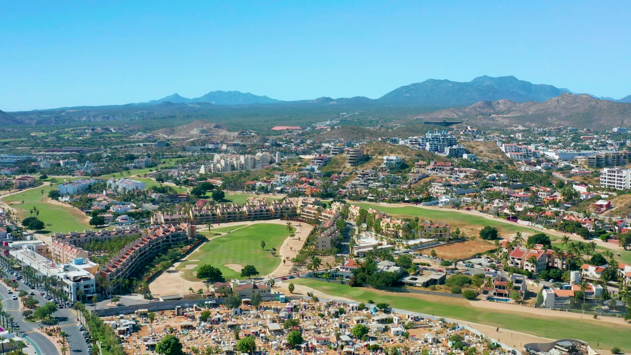 San Jose del Cabo aerial frame with clustered homes, a tan open area, green fairways, white buildings on the left, mountains on the horizon, and no visible people.