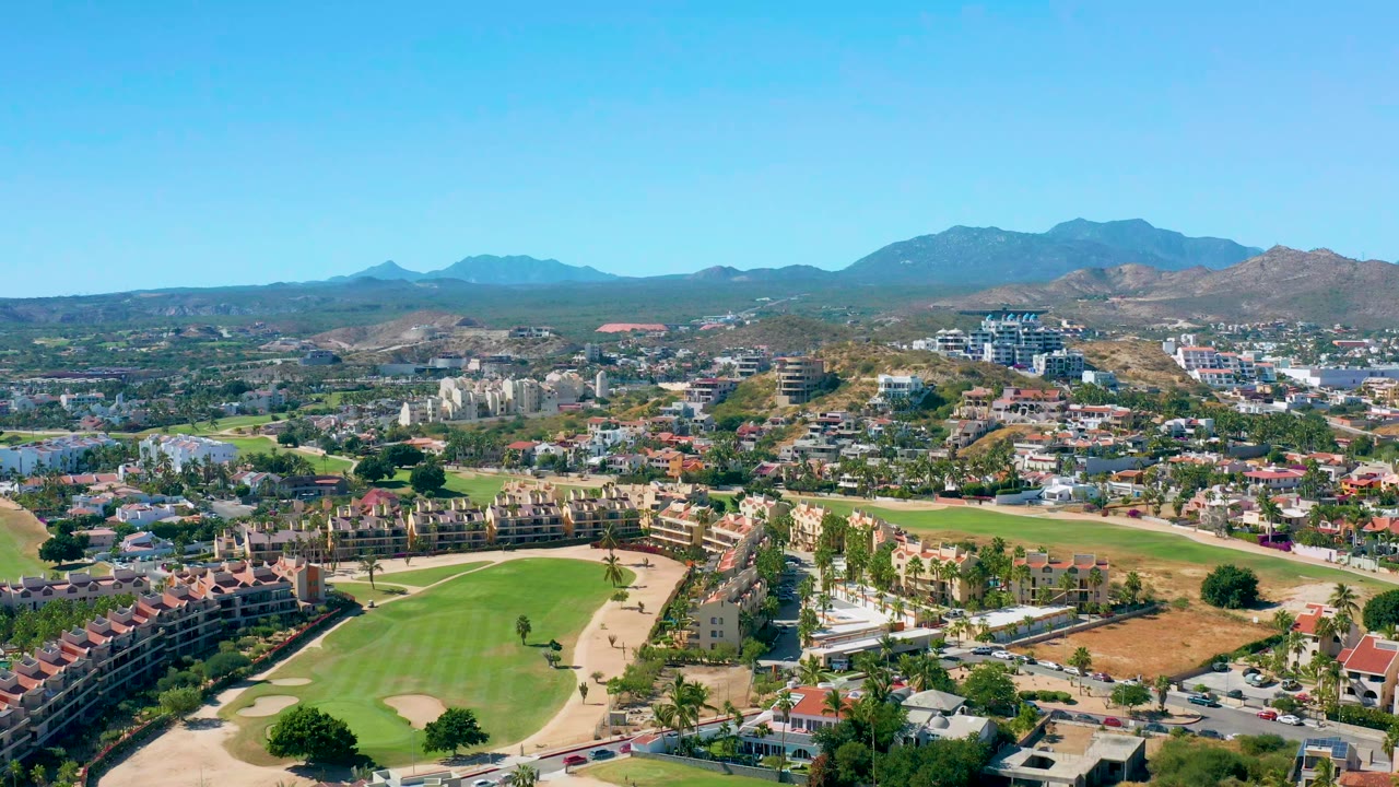 Opening aerial view over San Jose Del Cabo with golf fairways, condo rows, low homes, dry hills, distant mountains, and no visible people.