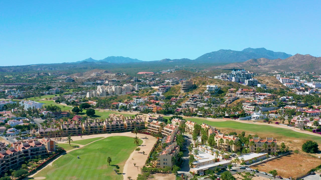 Opening aerial frame of San Jose del Cabo showing a green golf course in front of red-roof homes, palm trees, dry hills, distant mountains, and no visible people.
