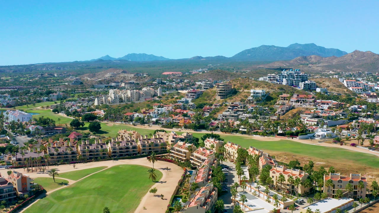Opening aerial frame over San Jose del Cabo with green golf fairways in the foreground, red-roof homes and low buildings in the middle distance, mountains on the horizon, and no visible people.