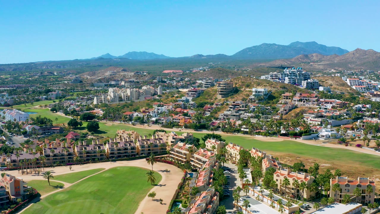 Elevated San Jose del Cabo frame with apartment-style buildings and red-roof homes near the lower edge, golf greens through the center, dry hills in back, distant mountains, and no visible people.