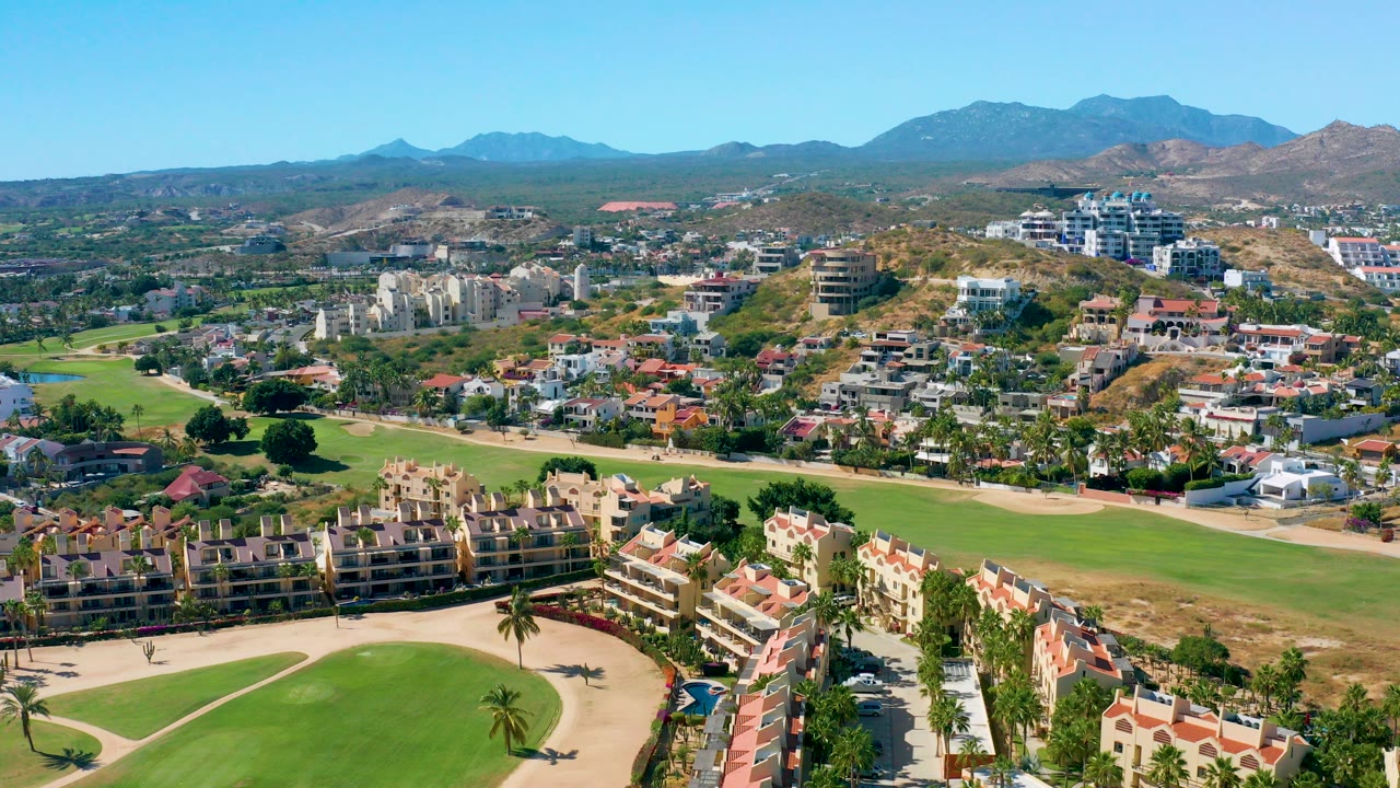 Early aerial frame over San Jose Del Cabo showing golf fairways, beige residential buildings, palms, roads, dry hills, distant mountains, blue sky, and no visible people.