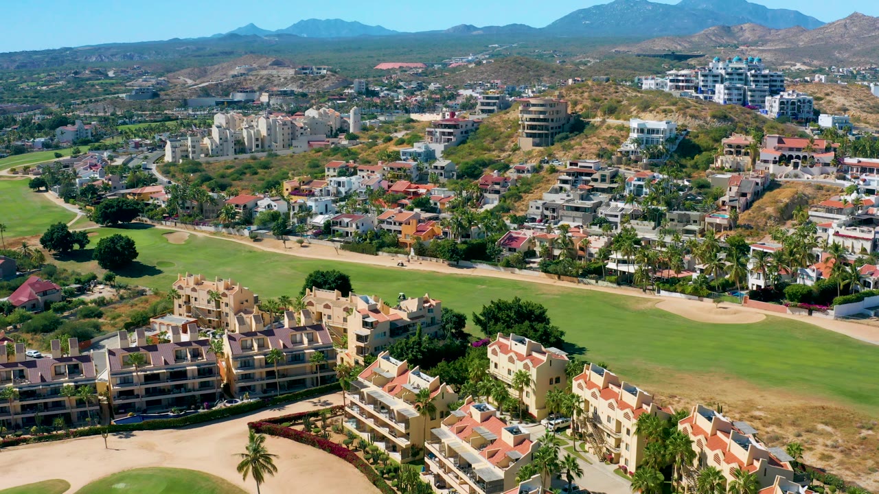 Mid-pan aerial over San Jose del Cabo with foreground condos, a long green fairway, sand traps, hillside residential development, mountains, and no visible people.