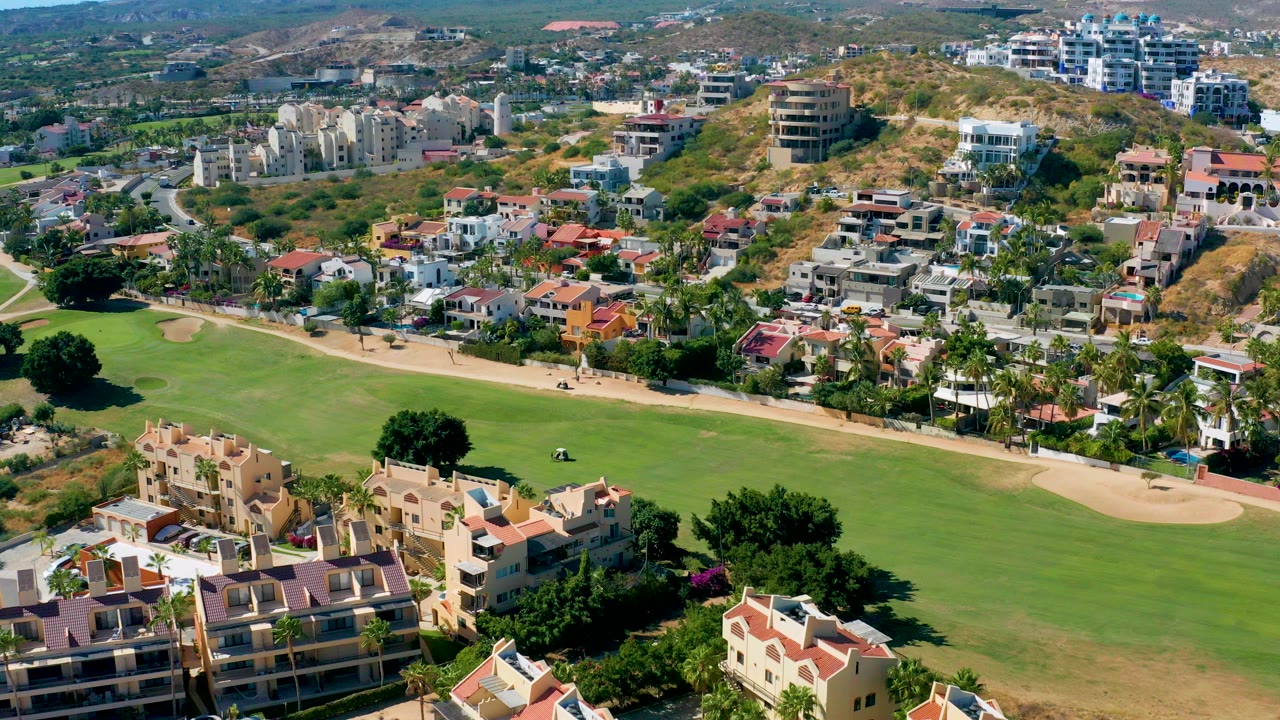 Aerial frame in San Jose Del Cabo showing a broad green fairway, tan cart path, apartment roofs in the foreground, hillside residences beyond, and no visible people.