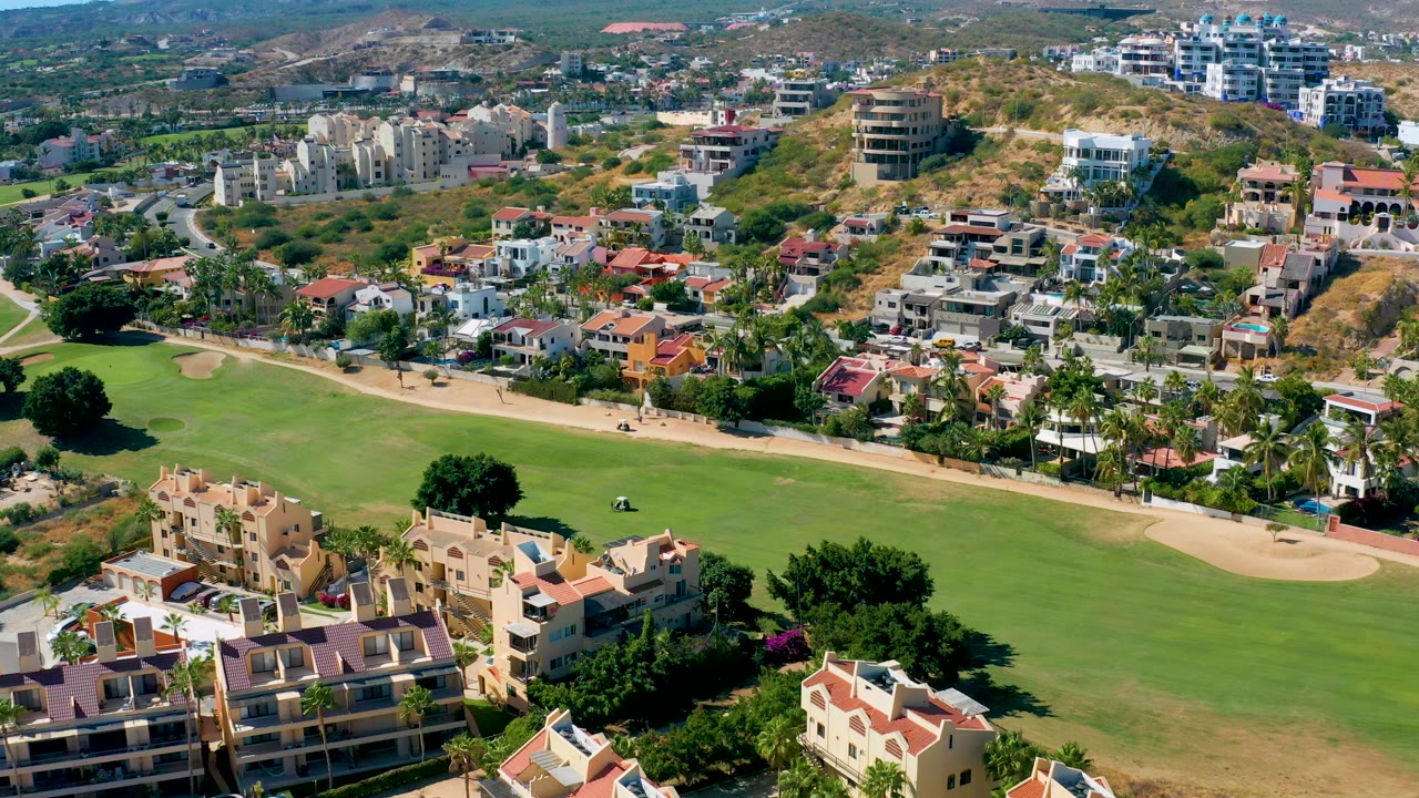 Elevated San Jose Del Cabo view with the green fairway across the center, a tan path cutting along the homes, foreground roofs, hillside buildings, and no visible people.