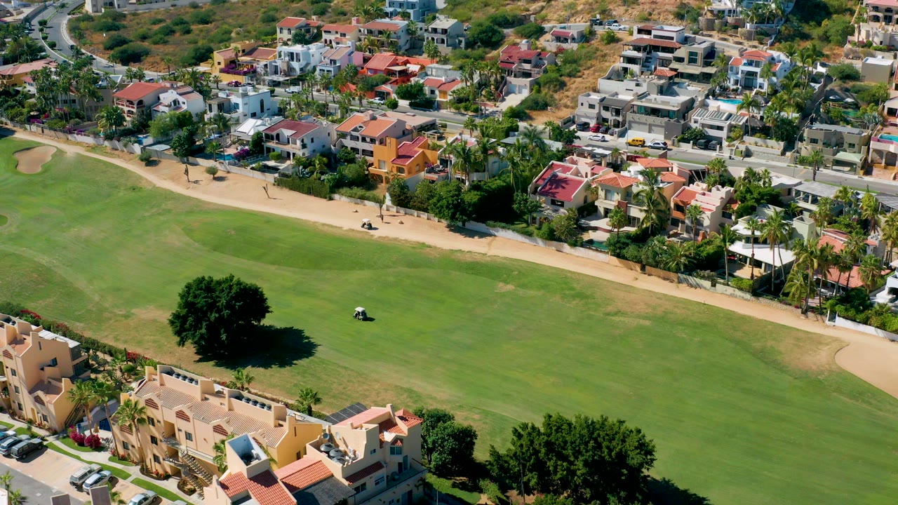 Aerial frame from San Jose Del Cabo, Baja California Sur showing a small golf cart on a green field below residential homes and roads, with no visible people.