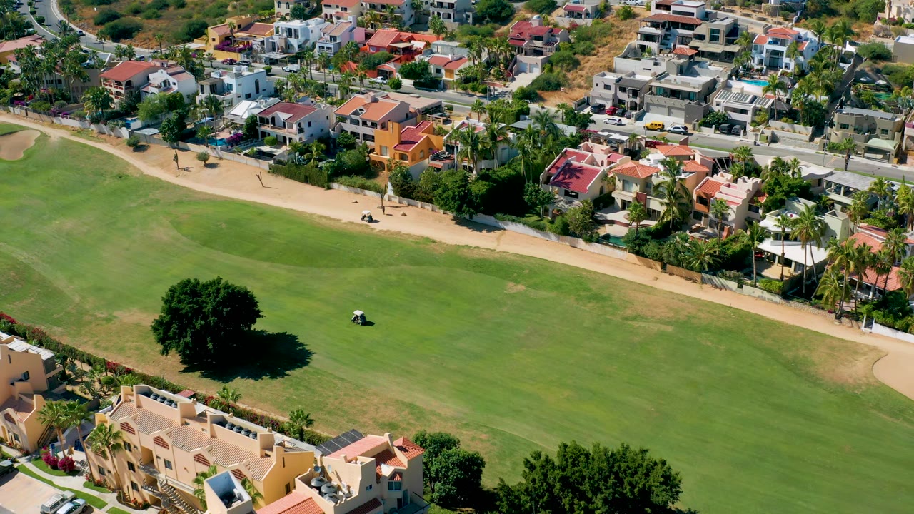 Drone-height frame from San Jose Del Cabo showing a golf cart near the center of the grass, a curving tan path, and rows of homes beyond the field, with no visible people.