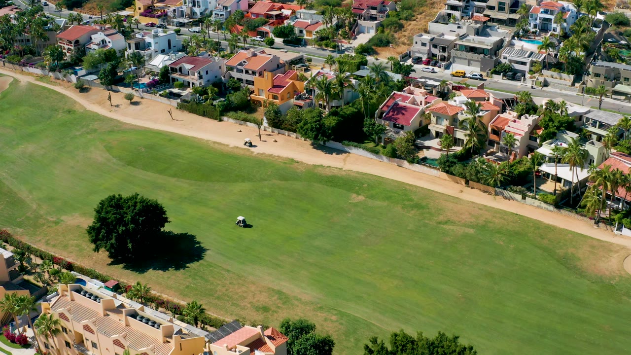 Aerial frame with a golf cart on grass and colorful homes beyond a path.