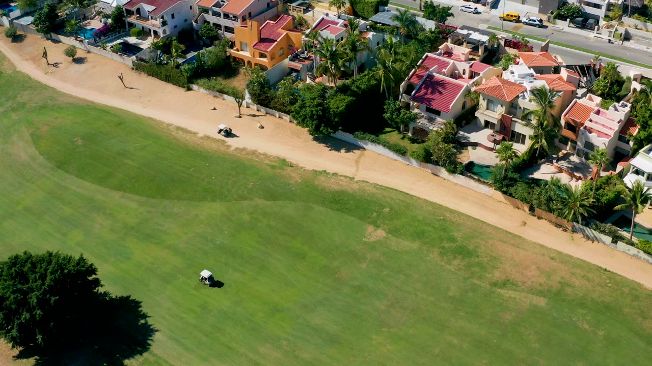 Aerial frame in San Jose del Cabo shows a small white golf cart on the golf course below a sandy path, with red-roof homes, palms, and no visible people.