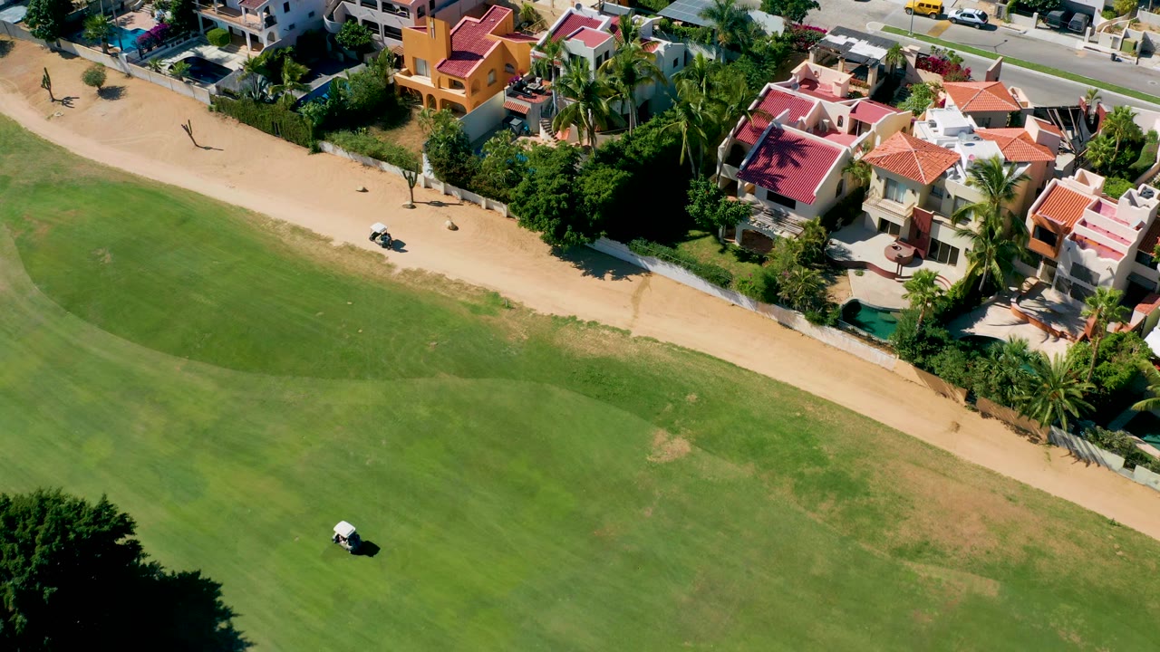 The cart appears lower in the frame on the green course, with a wide sandy path, roadside homes, palms, and no visible people in San Jose del Cabo.