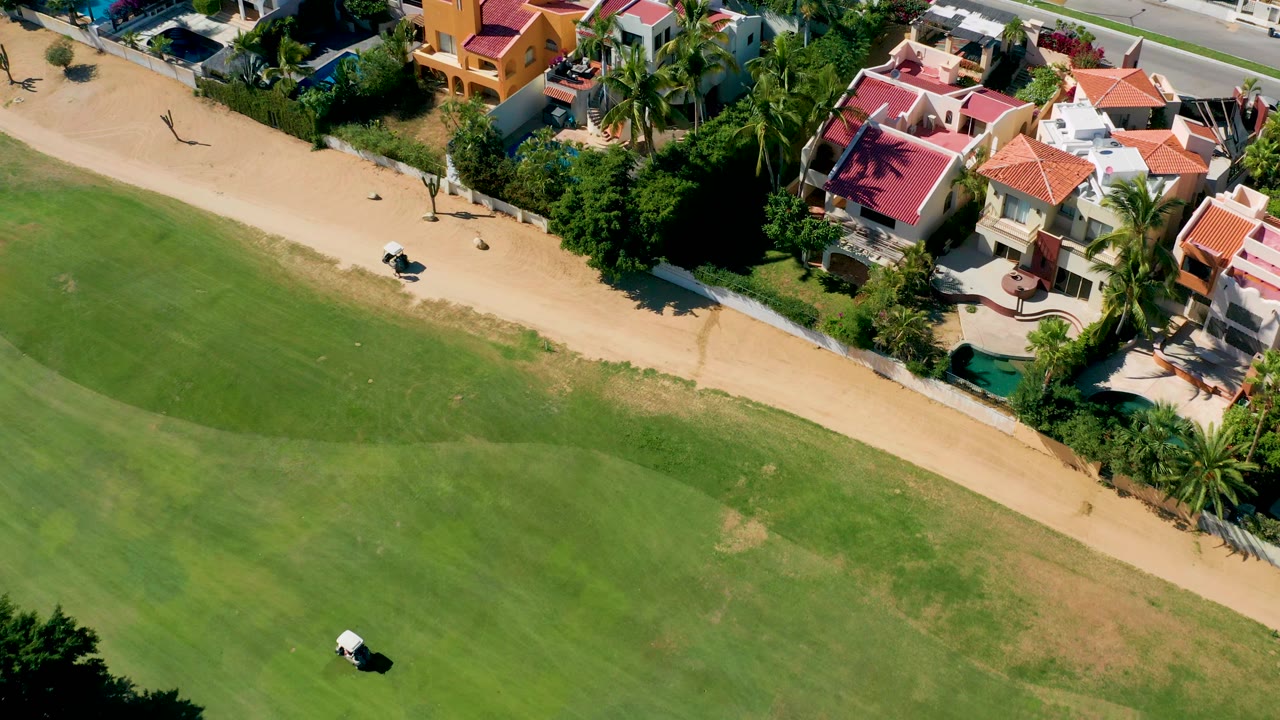 Golf cart on the edge of the green near a sandy path and houses with pools.