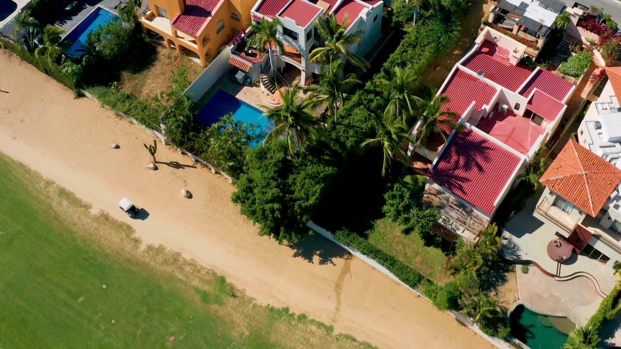 Overhead San Jose del Cabo frame with a sandy road and grass along the lower left, red-roofed homes, blue pools, palm trees, and no visible people.