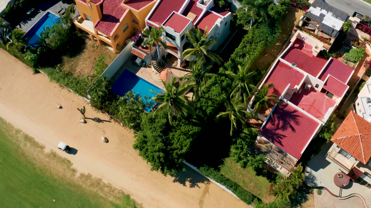 Top-down San Jose del Cabo frame showing the sandy road near the lower left edge, clustered red roofs, bright blue pools, palms, and no visible people.