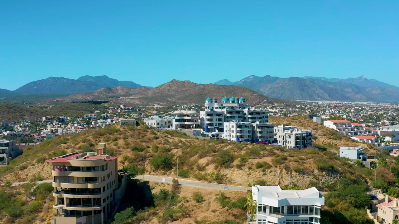 Early frame with the rounded foreground building lower left, white hillside apartments near center, San Jose del Cabo city beyond, mountains, and no visible people.