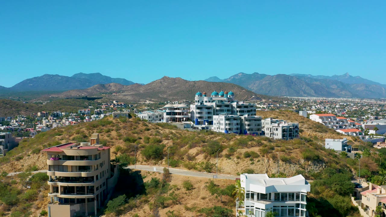 Aerial frame showing dry San Jose del Cabo slopes, clustered white residential buildings, a distant urban area, layered mountains, and no visible people.